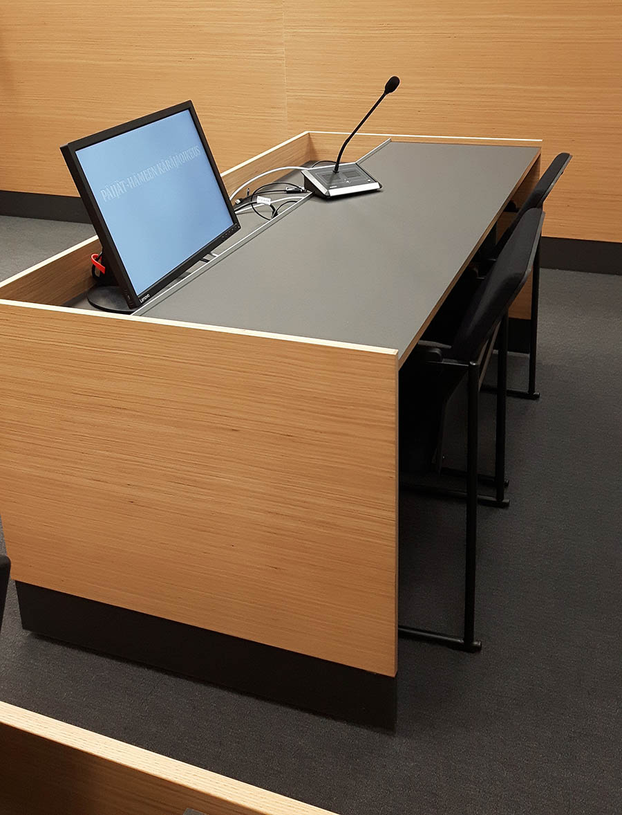 Kuvassa on pöytä ja sen edessä tyhjä tuoli. Pöydällä on tietokoneen näyttö ja mikrofoni. 